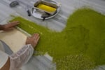 A baker rolls chopped pistachios into baklava dough at the Celebiogullari factory. There is a large pile of chopped, green pistachios along with a tray of melted butter.