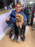 A woman in a blue shirt in a wheelchair with a small brown dog on her lap.