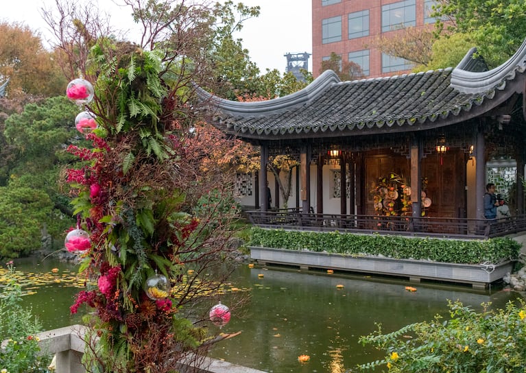 A floral arrangement stands in front of a lake and a pavilion inside a garden.