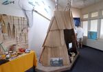 A replica teepee sits on a wooden platform in the museum right next to a table and display of tribal regalia and other tribal artifacts. The wall in the back has salmon decorations alongside a fishing net.