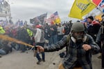 Rioters assault police protecting the U.S. Capitol on January 6, 2021. A man in the foreground is spraying an orange liquid. Crowds of people are packed together. Some are holding up flags, including the U.S. flag, "Don't Tread on Me" flags, Trump flags and a Confederate flag.