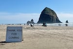 On a beach, a white sandwich board is printed with "THANK YOU FOR COMING." The ocean and Haystack Rock are in view in the background, with a sunny blue sky.