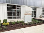 A large white and gray building with big windows has a sign in front of the entrance that says "Farmers Market Pavilion."