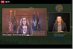 An image of a computer screen shows Oregon Gov. Kate Brown speaking in front of flags on the left of the screen, while a man in a separate window to the right provides sign-language interpretation.