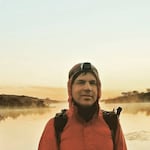 A man wearing a red jacket, backpack and headlamp looks at the camera with a landscape in the background. Only his upper torso is visible.