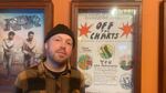 Eric Isaacson stands next to the hand-drawn event poster for the Off the Charts screening and song contest in the lobby of the Hollywood Theatre. He's wearing a yellow and black plaid flannel and a black beanie.