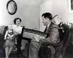 A black-and-white photograph of a man sitting in a sofa chair with a large records book and a young boy leaning over his shoulder. The man is interviewing a woman who has an infant in her lap.