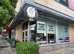 A close up of a black-and-white round sign on the corner of a building. The sign says "doe" and features a deer inside of a donut. The windows of the shop say "black lives matter" on them. A bush is visible in the foreground.