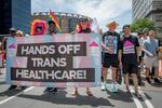 Several people carry a large banner that says "Hands off trans healthcare" as they walk down a city street. Another person has a small sign that says the same thing.