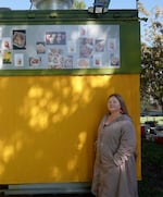 Woods stands in front of her bright yellow and green food cart as sunlight filters through the trees. Photos of her menu are posted at the top of the cart.
