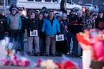 Community members stand shoulder to shoulder, some holding hands, at a vigil. There are bouquets of flowers on the ground.