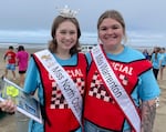 Two smiling girls, wearing official judges vests, sashes with their titles, and blue event t-shirts. The girl on the left is wearing a tiara and holding a clipboard.