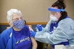 A health-care worker in full protective clothing administers a vaccine to a seated person wearing a mask.