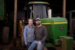 Amanda Hall stands next to a green tractor, and Reid Hall sits on the tractor's front wheel, with his arm around Amanda Hall's waist.