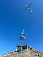 A helicopter carefully drops building materials onto the base of the High Rock lookout to begin the reconstruction in 2025. The lookout sits atop a 5,685 foot sharp peak, surrounded on three sides by sheer cliffs.