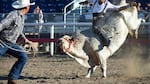 A man rides the back of a bull that is kicking up its hind legs as another man nearby holds a lasso.