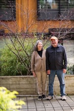 This photo shows Mary and Ray Johnston standing next to one another outdoors in front of a small tree. Ray Johnston is on the right side of the frame and is wearing blue jeans, a long-sleeved blue shirt and a yellow baseball cap. Mary Johnston is standing toward the middle of the frame and is wearing light brown pants, a black shirt and a light brown cardigan.