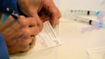 A hand holds a pen and fills in a vaccine card on a table where syringes sit.