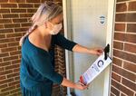 A woman with her hair tied in a single braid behind her back hangs an informational placard from a glass door's handle on a brick home's front porch.