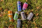 A variety of colorful cans of THC drinks are arranged on a grass. Some flavors include The Tenacious Seed's Blood Orange beverage, Silly Goose's root beer THC-infused soda and Mountain High Seltzer's Berry Wild.