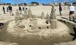 A sandcastle with half a dozen towers, connected by walls in a circle, and being breached on the left side by a sand-sculpted octopus. A moat surrounds the castle. Spectators walk by on the beach in the background.
