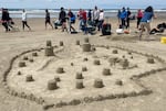 Over a dozen simple sandcastles of various sizes is surrounded by a wall made of sand. Spectators walk by in the background.