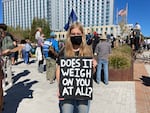 A young woman wearing a facemask looks into the camera while holding a sign with the message, "DOES IT WEIGH ON YOU AT ALL?" Behind her are several people and an office building.