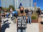 A young woman wearing a facemask looks into the camera while holding a sign with the message, "DOES IT WEIGH ON YOU AT ALL?" Behind her are several people and an office building.