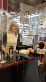 A female clerk with blond hair and glasses smiles behind the check-out counter at a video store. There is a clear plexiglass barrier separating the employees behind the counter from customers. In front of the counter, a customer is partially in view, checking out a stack of DVDs. On the counter sits a tip jar adorned with a picture of Elvira.