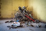 This photo shows a tangled pile of old, thick wires and metal pipe fittings sitting in the corner of a basement at a home. The basement has pale yellow cinder block walls and a gray concrete floor that's covered in dust and grit.