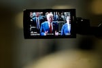 This photo shows former President Donald Trump on a camera monitor as he speaks to the press in February outside Manhattan Criminal Court. He's wearing a blue suit jacket, a red tie and a white shirt. Many people in suits and law enforcement uniforms stand behind him. 