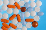 Macro photograph of various over-the-counter pain medicines, including Aleve, Tylenol, Advil, and various other NSAIDs and acetaminophen pills that are a variety of colors, including white, orange, brown, and light blue, and range from circles to elongated ovals, close up on a bright blue backdrop. 