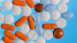 Macro photograph of various over-the-counter pain medicines, including Aleve, Tylenol, Advil, and various other NSAIDs and acetaminophen pills that are a variety of colors, including white, orange, brown, and light blue, and range from circles to elongated ovals, close up on a bright blue backdrop.