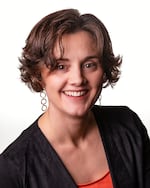 A close-up portrait of a woman smiling at the camera. She's wearing an orange shirt, a black sweater, and earrings.