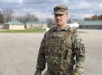 A person wearing military camouflage uniform looks off into the distance, with blurred gravel or pavement and buildings visible in the background.