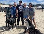 Five people pose smiling on the beach wearing gold medals. The man in the center wears an Elliott Smith t-shirt. Their three dogs are also pictured in the frame.