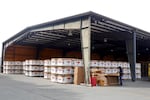 A large open air warehouse features tall piles of watermelon boxes as a worker builds more boxes.