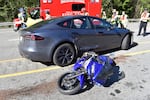 This photo shows a blue motorcycle lying on its side on a road. Behind it is a dark gray Tesla Model S sedan. Several first responders wearing helmets and orange and yellow reflective vests stand behind the Tesla. A red ambulance is parked in the background.