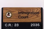 This file photo shows a rectangular sign that indicates the location of an immigration court in New York City. The sign says "Immigration Court" on a wood-patterned background, and the round Department of Justice seal, with an eagle on it, is toward the top-left corner. The bottom of the sign says "C.R.: 23" and "2035," with braille underneath.