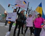 Four people hold protest signs and wave Ukrainian flags. One sign reads, "Ukraine deserves to stay free." Another reads, "Stop Putin NOW!!"