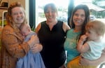 Three woman pose for a photo in an indoor setting. The woman on the left holds an infant while the woman on the right holds a toddler.