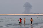 Three boys play in the shallow waters of the Strait of Hormuz, as a plume of smoke rises from an explosion in the background, off Bandar Abbas, Iran, Monday, July 13, 2026.