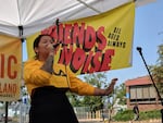 A person sings into a microphone while standing on an outside stage. A banner in the background reads "Friends of Noise All Ages Always"