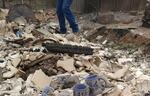 A person's jean-clad legs are visible as they walk through the ruble of a burned home. Three ash-covered coffee mugs are visible in the foreground amid a pile of broken drywall and charred debris.