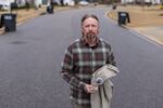 In this photo, Derek Copeland stands in a street holding a U.S. Department of Agriculture work shirt that has a round patch with a logo on it. Copeland has a beard and is wearing a plaid, button-down, long-sleeved shirt.