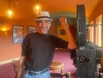 David Fox poses by an old movie projector in the lobby of the Hollywood Theatre. He's wearing blue jeans, a black t-shirt, and a tan and black fedora.