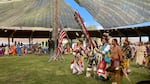 Four men march in a small, open-air arena. One man carries an American flag while another carries an Eagle Staff