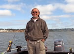 A man stands and smiles on a boat, with a fishing rode to his side and water in the background.