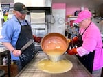 Two white male candy shop workers hold a kettle and pour taffy mixture onto a metal cooling table.