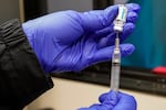 A gloved hand uses a needle to extract vaccine from a vial.